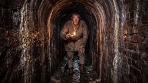 A medieval Guerchi holding an oil lamp inside the narrow underground tunnels of Bottini di Siena, surrounded by damp brick walls.